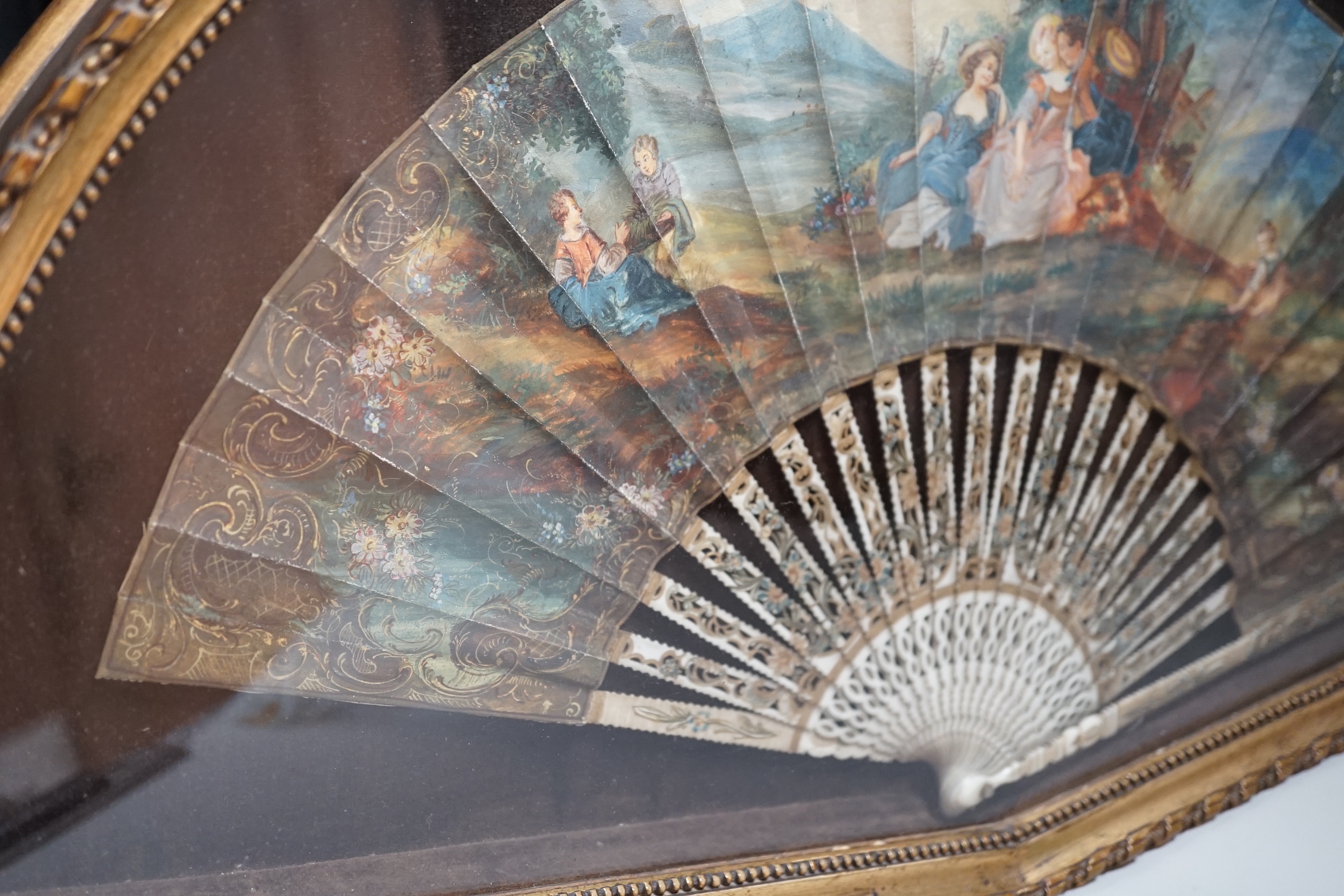 A 19th century French bone fan with pierced decoration, hand painted with figures wearing 18th century dress before a landscape, framed, the frame 62cm wide